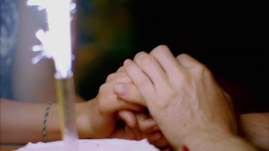 A man holds his girlfriends hand as sparks fly from a firework in a pink birthday cake in front of them