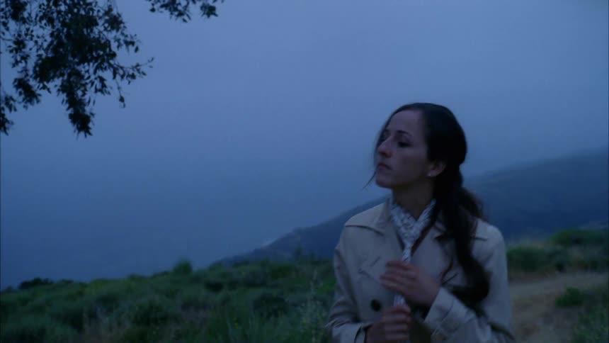 Woman with a black ponytail a brown trench coat walks on a dirt road and wears a scarf around her neck at dusk on a cloudy day