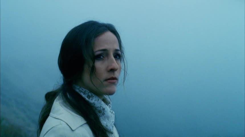 Woman with black wavy hair and a brown trench coat walks uphill and looks to the right and then smiles slightly in the foggy mountains