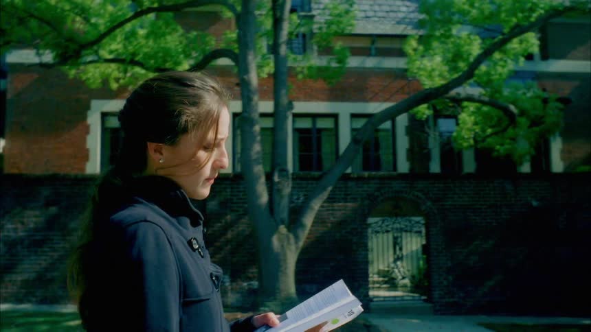 Woman with a ponytail and a black overcoat walks on a pavement while reading a book