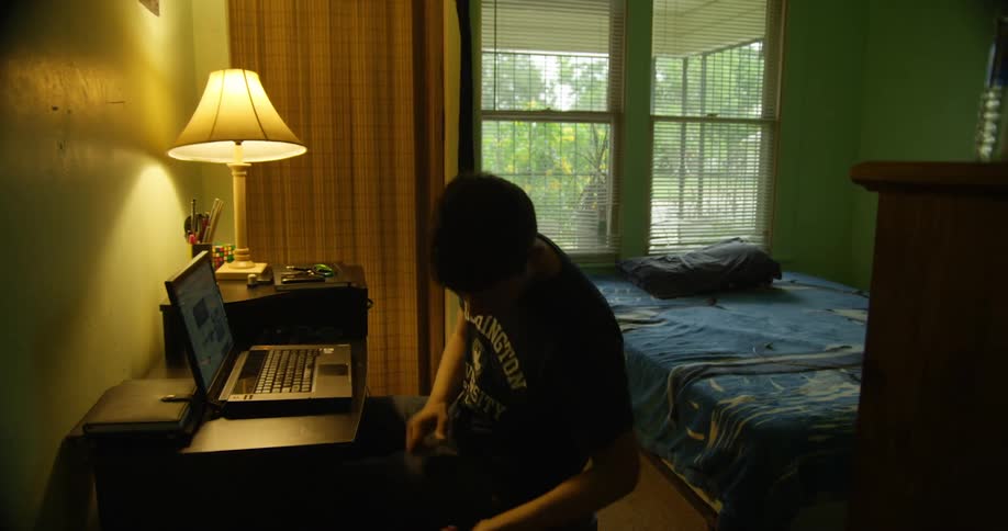 A young man sitting in front of a laptop at a desk reaches to retrieve the charging cable for the laptop and plugs it in
