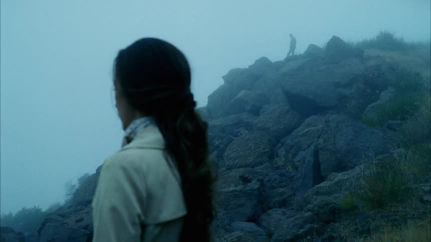 A woman stands on a foggy mountain looking out over the terrain as male figure shrouded in fog starts to approach her from higher on the mountain