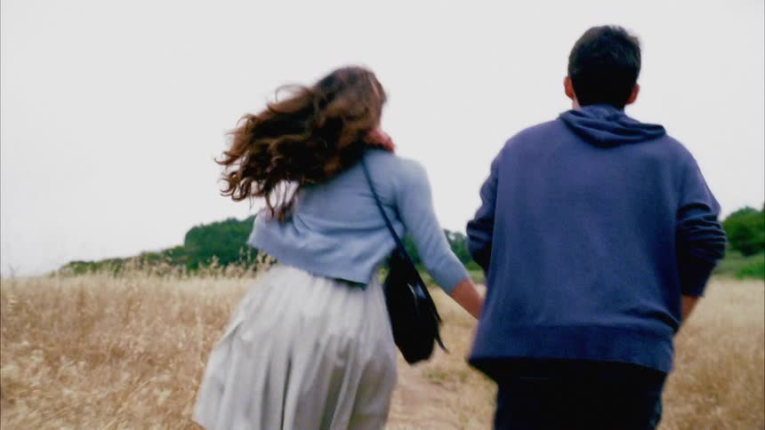 Couple of teenagers holding hands run through a field of dry grass
