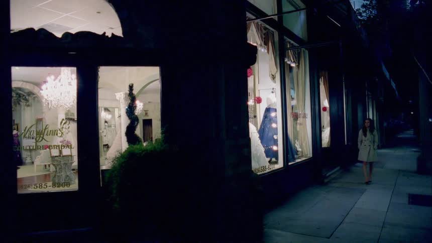 A woman in a trench coat walks down a sidewalk at night before stopping to look into the window of a bridal shop