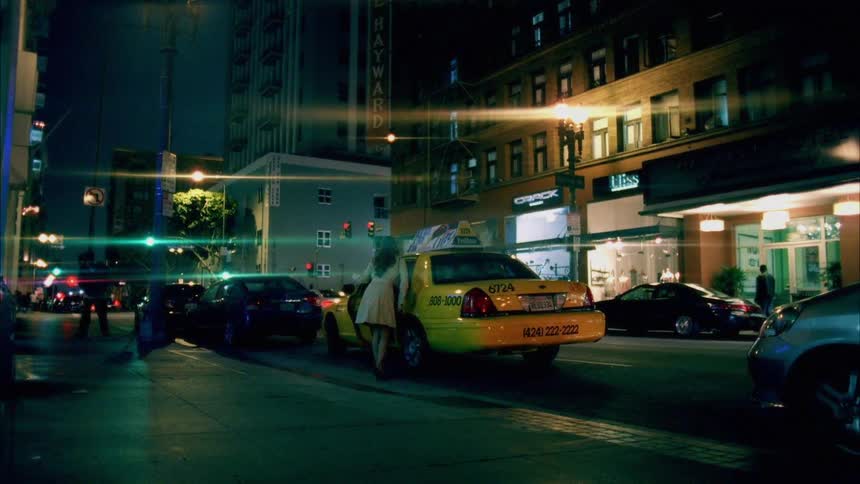 A woman in a trench coat enters the backseat of a taxi cab waiting at the side of the road in a metropolis at night