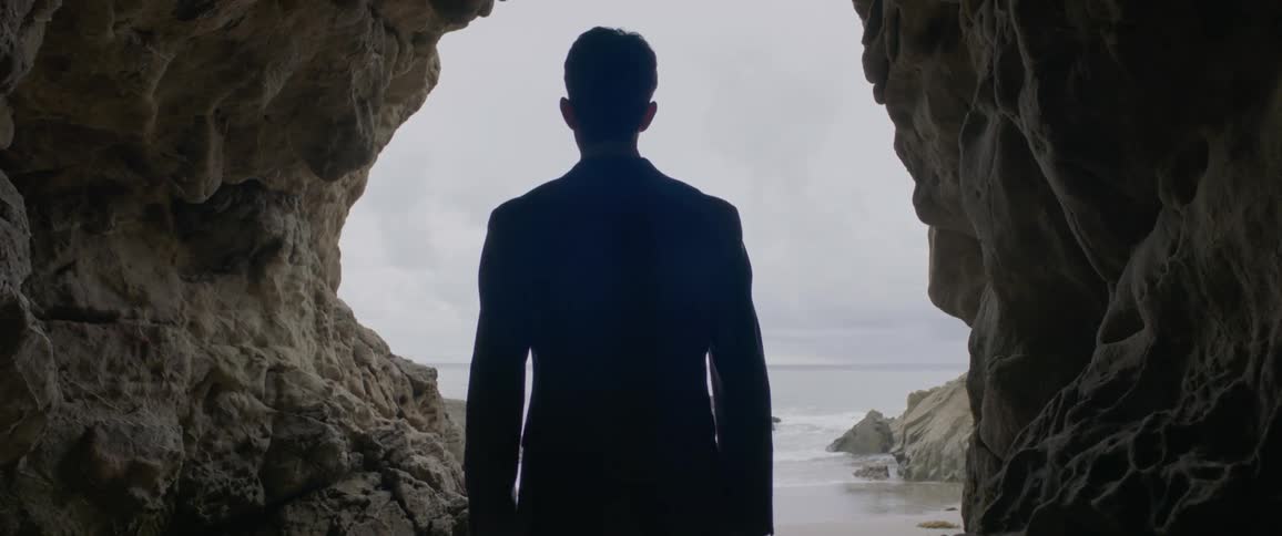 A man is standing at the entrance to a small cave looking out over the water