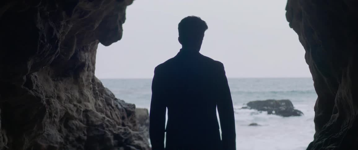 A man stands inside the opening of a cave overlooking the ocean