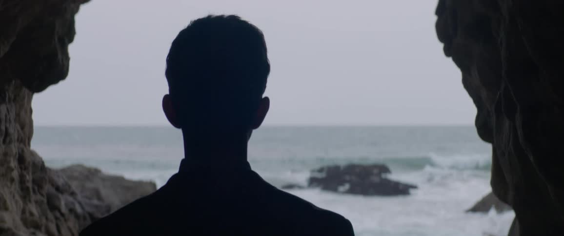 A man standing at the entrance to a cave is looking out over the ocean