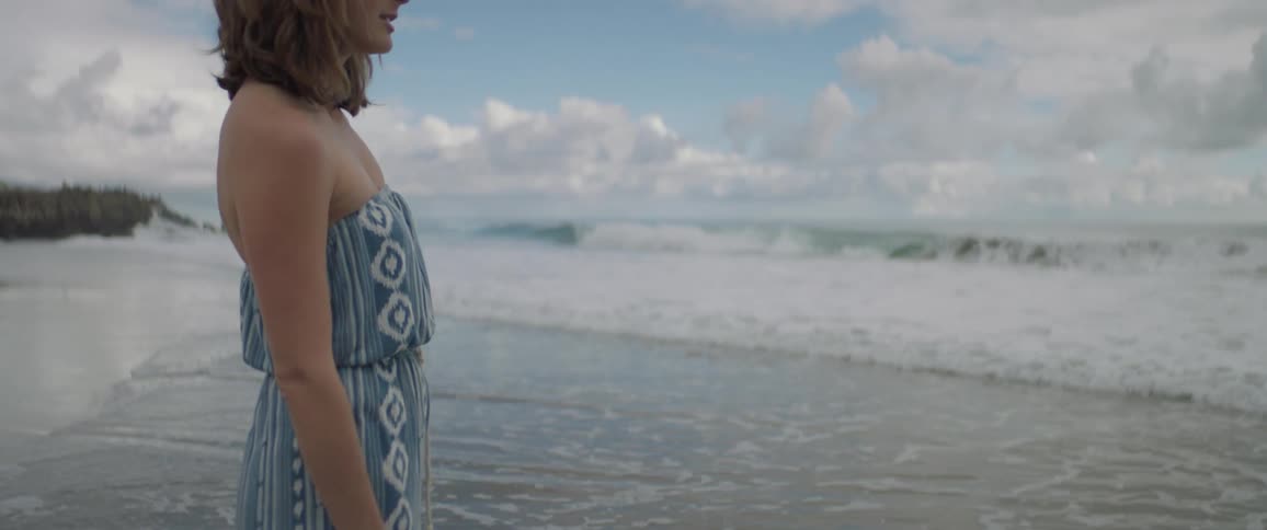 Waves hit a woman's feet as she stands at the beach facing the water