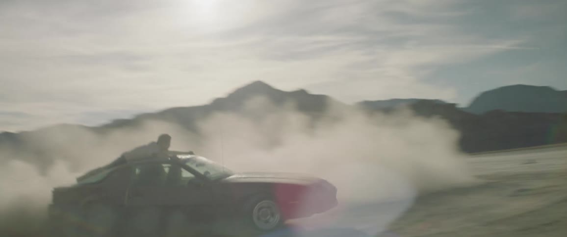 A man hangs on the roof of a car as it does donuts
