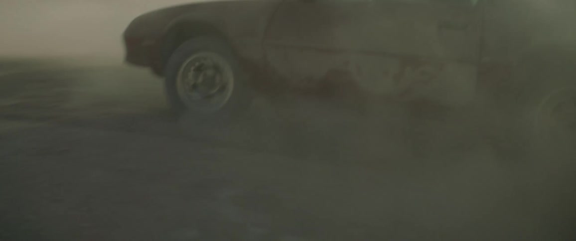 Woman driving erratically in a dusty area and enjoying it while a man clings to the top of the car near sunset