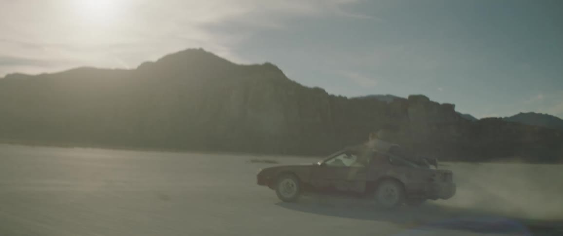 Woman driving erratically in a dusty area and enjoying it while a man clings to the top of the car near sunset