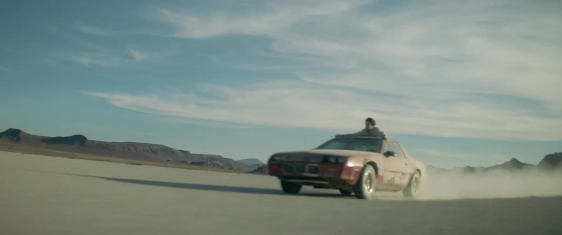 Driving in a dusty area while a man clings to the top of the car