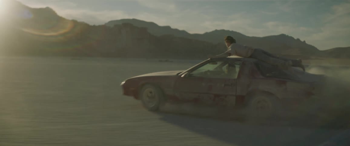 Woman driving erratically in a dusty area and enjoying it while a man clings to the top of the car on a cloudy day