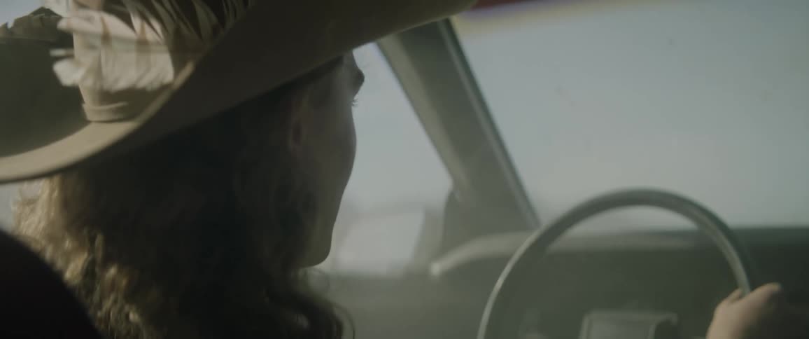 A person takes off their cowboy hat as they drive a car in a dusty environment during the day