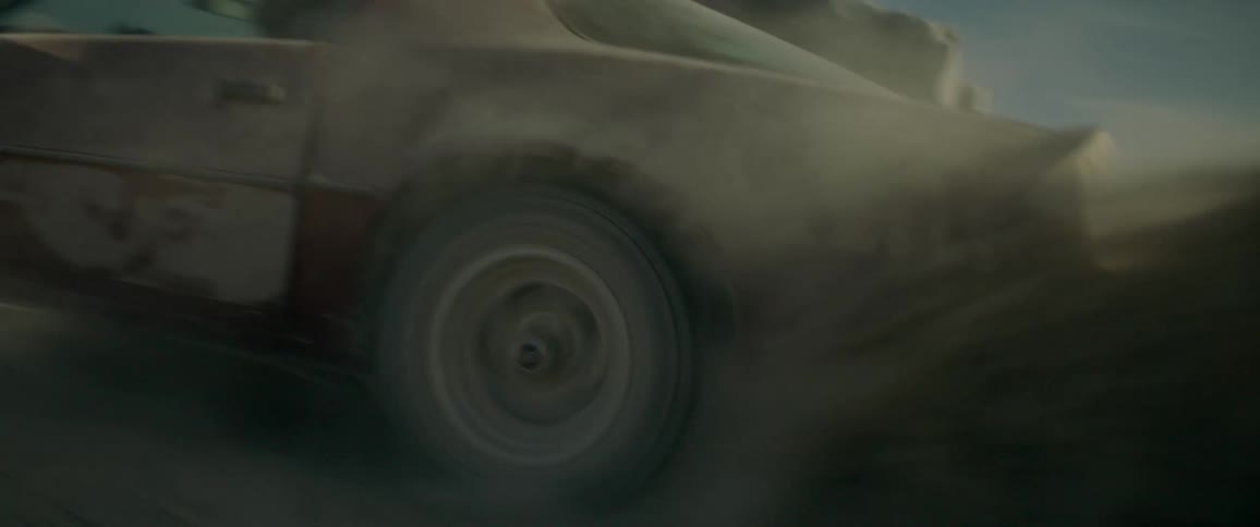 A red car does donuts in the dirt as a man holds onto the roof