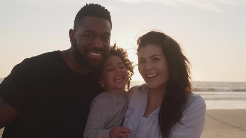 Black father son and white mother smiles and pose for the camera on the sand beach at sunset next to the ocean