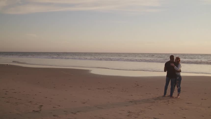 Couple hugs lovingly standing on the sand beach at sunset next to the ocean
