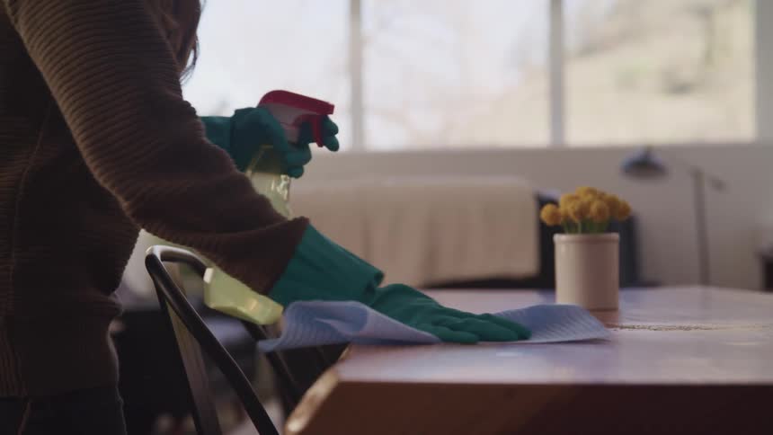 A woman in rubber gloves sprays cleaning fluid onto a wooden table and begins wiping it with a cloth