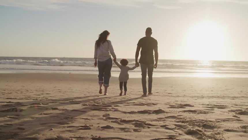 A mother and father hold their childs hand as they walk down the beach