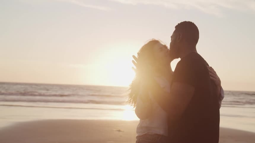 Black man and white woman dance on the sand beach at sunset next to the ocean