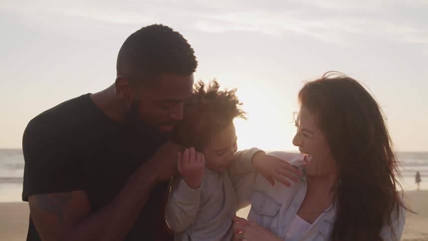 Black father and white mother hold toddler boy and tickle him on the beach at sunset next to the sea