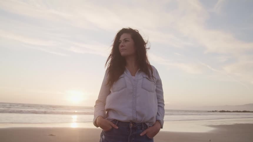 Woman with wavy hair a blue shirt and and hands in pockets looks to the right on the beach next to the sea at sunset