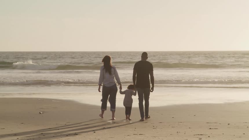 Black father son and white mother hold hands and walk barefoot on the beach next to the ocean at sunset