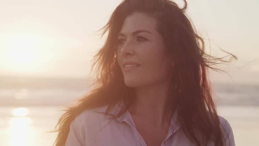 A woman is smiling outdoors by the coast while the sun is setting