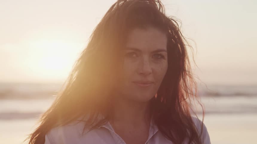 Woman with black wavy hair faces the camera with eye contact on the sand beach next to the sea at sunset