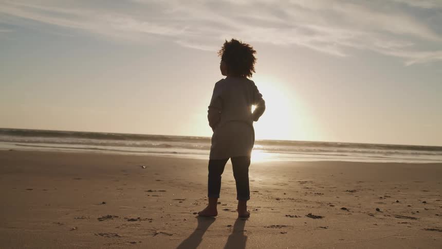 boy walking and turning around on the sand beach next to the sea is silhouetted against the sunset