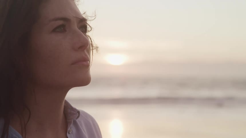 A middle aged woman stands on a beach as the sun descends behind the horizon