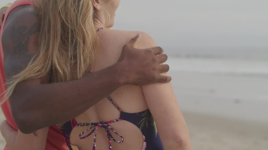 A man wraps his arm around a woman as they stand on the beach watching the ocean water