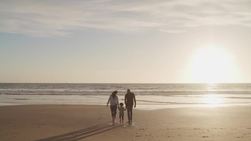 Family walk holding hands over the sands next to the shoreline of a beach at sunset