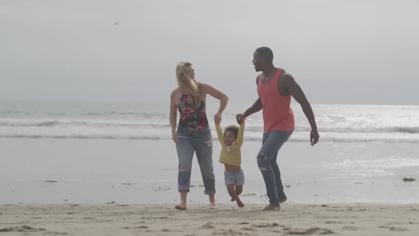 A man and woman hold their daughters hand and swing her in the air as they walk along the beach