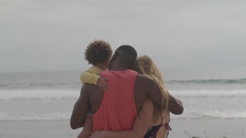 A man holds his child in his arm and rubs his wife arm as they stand on the beach and look at the water