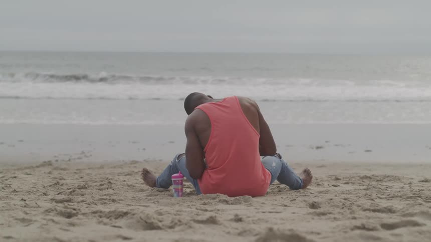 A barefoot black man sits in the sand of a beach while facing the ocean on a cloudy day