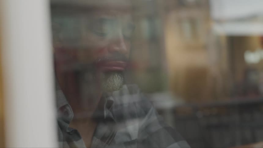 Bearded man behind the glass window of a store looks outside pensively