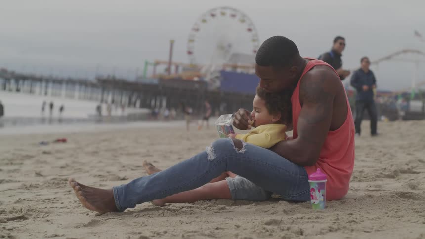 A man and his young daughter sit closely on a beach together eating a snack
