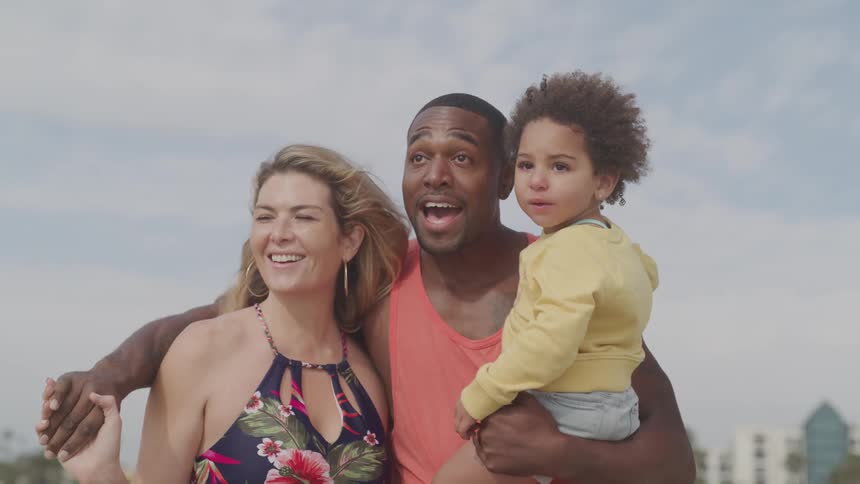 Father hugs mom and kisses her while carrying their child on cloudy day