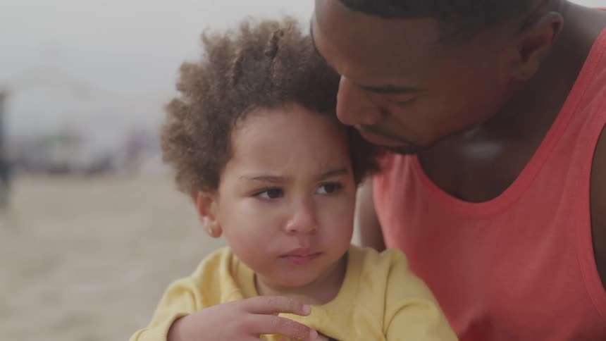 A man sitting on the beach kisses the forehead of a little girl sitting in his laptop eating candy
