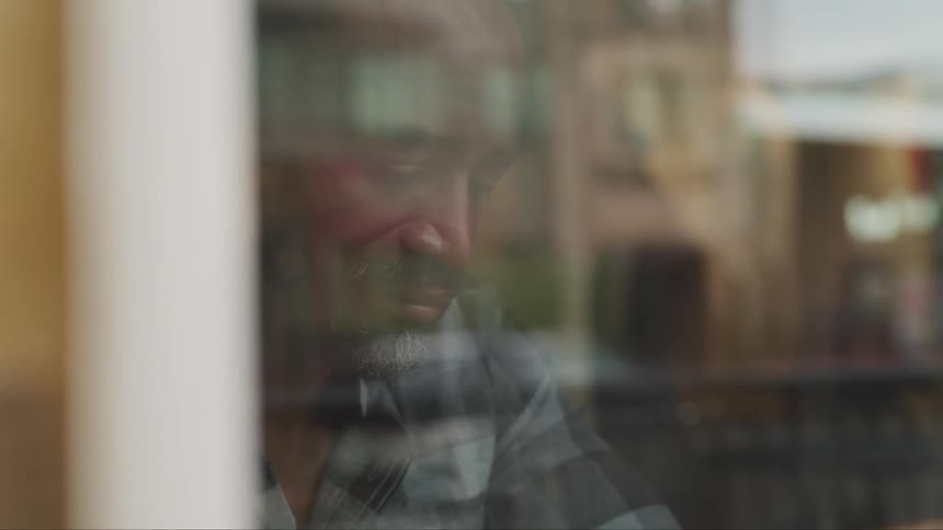 Black man with a beard and a checkered shirt drinks a glass of coffee at a cafe