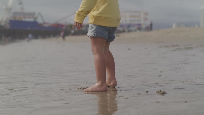 Young boy in jean shorts walks barefoot on the wet sand of a beach near a shipping freighter