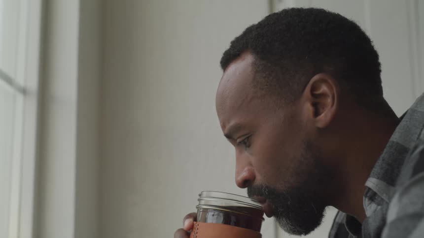 A middle aged black man drinks from a jar while sitting in front of a window