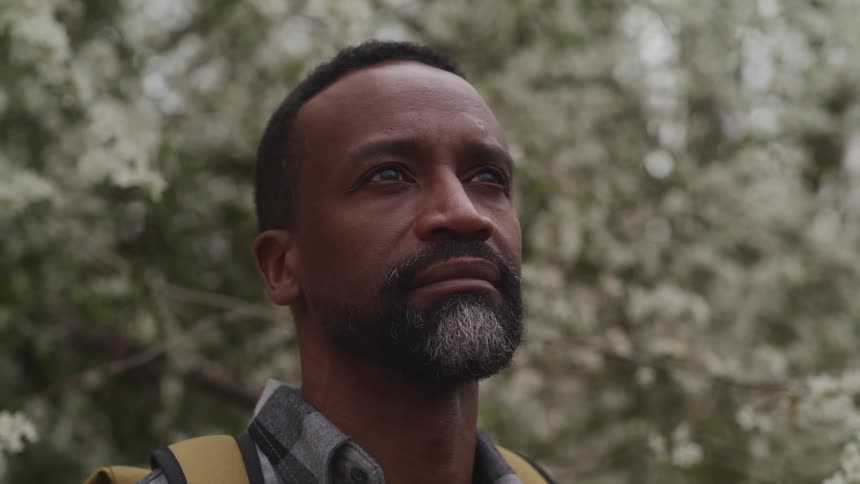 Black man with a beard yellow backpack and a checkered shirt looks up at the sky