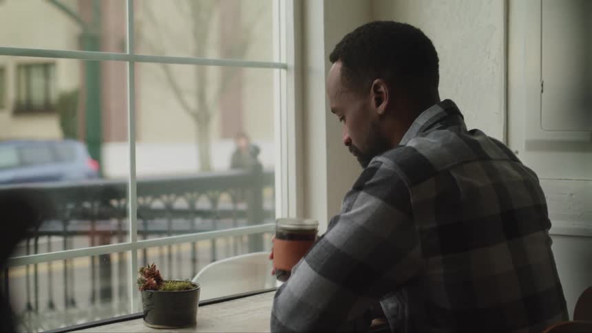 Young bearded man drinks coffee on a restaurant table while looking pensively out of a window