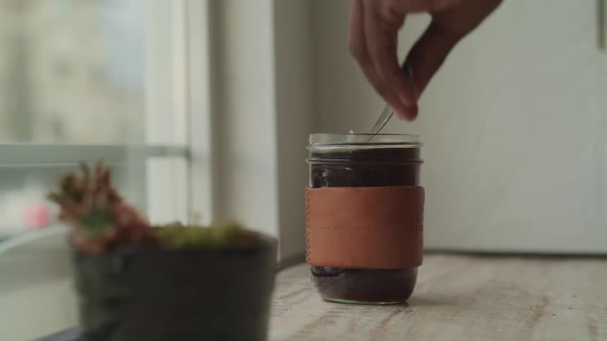 A hand stirs a jar of steaming hot coffee next to a window and potted plant