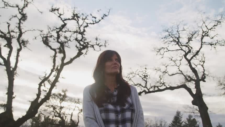A woman standing in front of a forest looks off in to the distance
