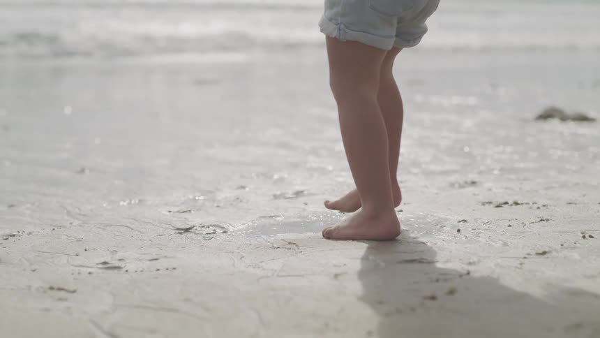 A child in denim shorts stands barefoot in the damp soil on a beach
