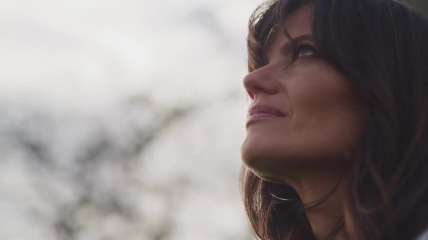 Side view of a woman with wavy brown hair and makeup looking up at the sky on a cloudy day